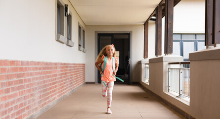 Full length of smiling caucasian elementary schoolgirl with backpack running in school corridor