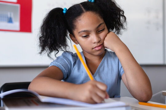 Biracial elementary schoolgirl writing on book at desk in classroom