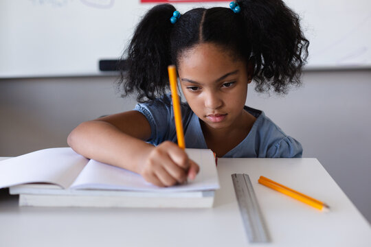 Biracial elementary schoolgirl writing on book at desk while studying in classroom