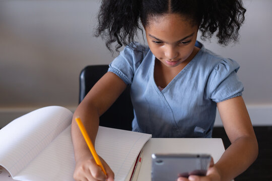 High angle view of biracial elementary schoolgirl using digital tablet while writing on book at desk