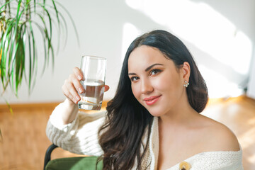 A young beautiful dark-haired woman in a knitted cardigan drinks clean water from a glass. Cozy house model portrait. Health.
