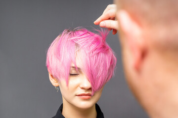 Hairdresser with hands is checking out and fixing the short pink hairstyle of the young white woman...