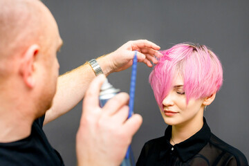 The hairdresser is using hair spray to fix the short pink hairstyle of the young caucasian woman in...