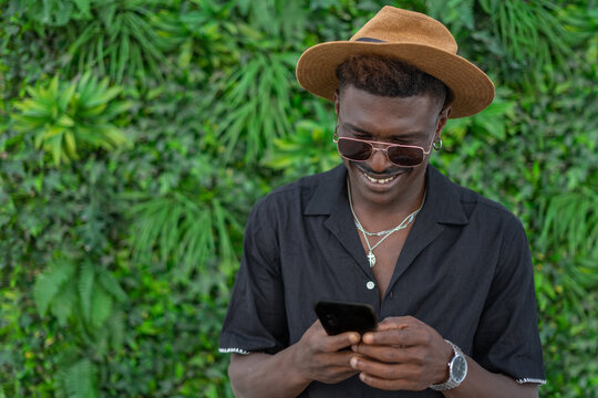 Chico negro con gafas de sol y sombrero, mandando un mensaje, con expresi&oacute;n de felicidad, en fondo con plantas verdes. Foto horizontal con espacio para texto.