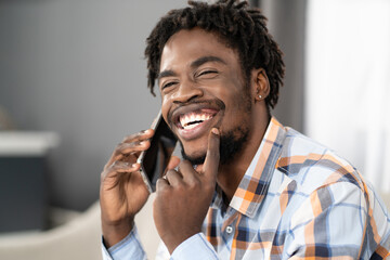 Happy smiling young African American man talking on the phone holding smartphone in hand looking away. Portrait of happy man having present conversation sitting on the sofa. Social media concept