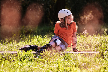 A tired child from sports is resting while sitting on the sidewalk on a summer day. Happy girl dressed in fall protection and roller skates sits on the track in the park. The young sportswoman  - obrazy, fototapety, plakaty