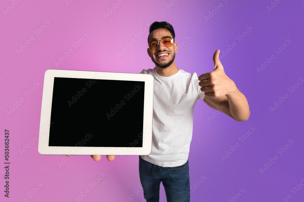 Poster happy young arab guy showing tablet pc with empty screen, gesturing thumb up, recommending new app o