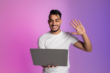 Cheerful young Arab guy waving at laptop, having video chat on webcam, conferencing online in neon light
