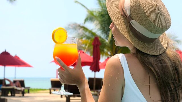 Beautiful girl in swimwear drinking colorful yummy cocktail at swimming pool. Woman in white swimsuit drinking non-alcoholic orange cocktail, relax in pool water, enjoy vacation at luxury hotel