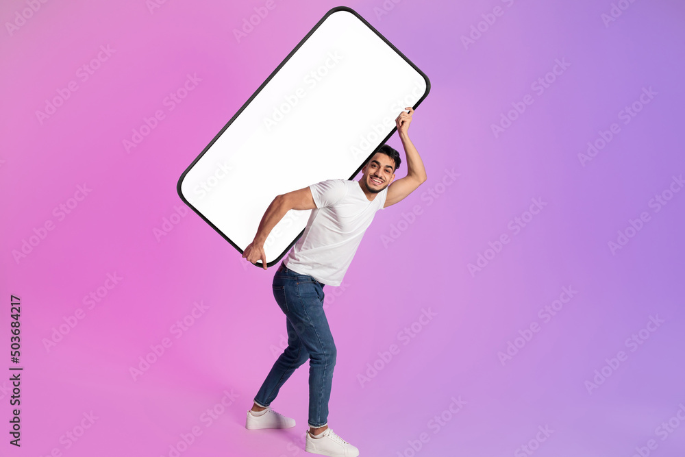 Wall mural Full length portrait of cheery young Arab man carrying huge heavy mobile phone with empty screen in neon light, mockup