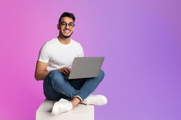 Young Arab man in casual wear working online, sitting cross legged and using laptop in neon light, full length