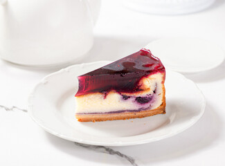 A piece of cheesecake with a berry layer and berry jelly on a light marble table among white dishes