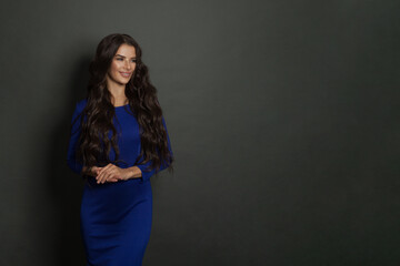 Beautiful young lady with long healthy hair and cute dress posing on dark gray background with copy space. Attractive brunette woman smiling