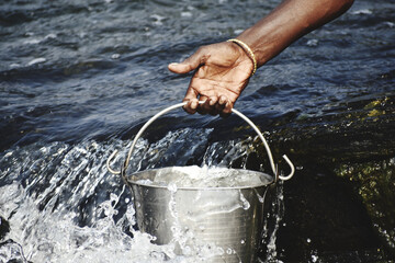 Closeup shot of a man's hand, taking a water from a river with a metal bucket © Ravindra Kumar/Wirestock Creators