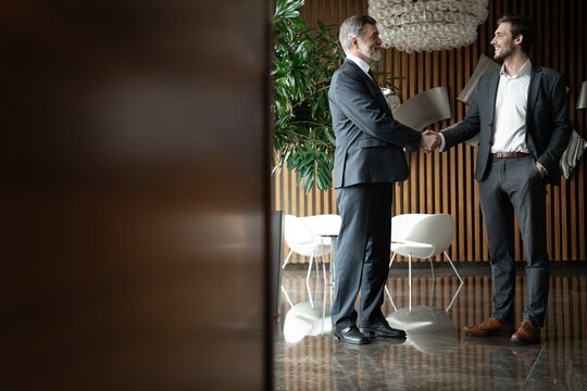 Two smiling businessmen shaking hands together while standing by windows in an office boardroom.