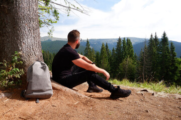 A young man in black clothes and a backpack sits alone outdoors with mountains and a forest in the background.