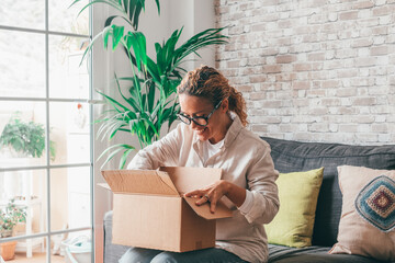 Overjoyed woman seated on sofa hold on lap small cardboard box open parcel client feels satisfied bought goods in internet, happy addressee received package from friend, quick delivery service concept - obrazy, fototapety, plakaty