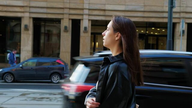 Confident woman walking down street. Side view. Beautiful career girl in leather jacket going around city. Slender long-haired woman in hurry on business. Entrepreneur lady