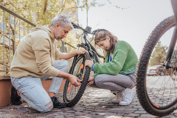 Happy father with teenage daughter repairing bicycle in street in town.