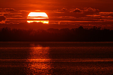 Sole rosso al tramonto con riflesso e luccichio sul mare e nuvole in lontananza - 503690692