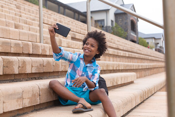 Smiling african american elementary schoolboy taking selfie while sitting on school building steps