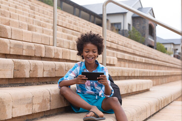 Smiling african american elementary schoolboy using smart phone while sitting on school steps