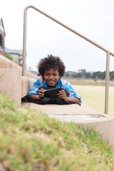 Portrait of smiling african american elementary schoolboy with smartphone lying on school steps