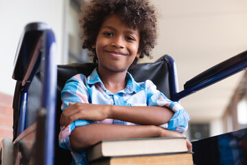 Low angle portrait of smiling african american elementary schoolboy with books sitting on wheelchair