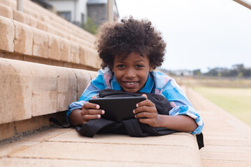 Portrait of smiling african american elementary schoolboy using smartphone while lying on steps
