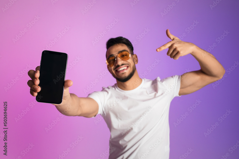 Poster smiling young arab man in sunglasses pointing at smartphone with empty screen in neon light, selecti