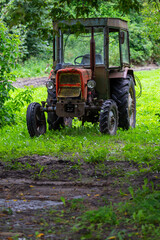 An old red tractor standing in the shade among the trees. The photo was taken on a cloudy day with good lighting conditions.