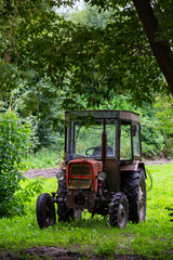 An old red tractor standing in the shade among the trees. The photo was taken on a cloudy day with good lighting conditions.