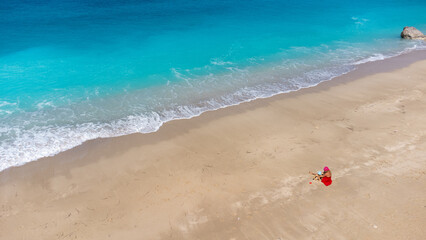 Fototapeta na wymiar Aerial view of woman in red dress sitting on the sandy beach with a baby, enjoying soft turquoise ocean wave. Tropical sea in summer season on Megali Petra beach on Lefkada island.