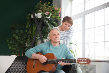 Fototapeta premium elderly couple spend time together. healthy lifestyle. emotional health. rest from gadgets. an elderly man plays guitar for his wife sitting on the couch at home. Slow life. Enjoying the little things