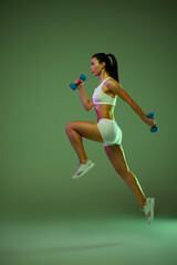 Full-length portrait of young muscular girl training, jumping with dumbbells isolated over green background in neon light