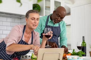 Schilderijen op glas Senior multiracial couple with wine watching online recipe and preparing food in kitchen at home © WavebreakMediaMicro