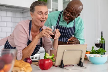 Fotobehang Happy senior multiracial couple with wine watching online recipe and preparing food at home © wavebreak3