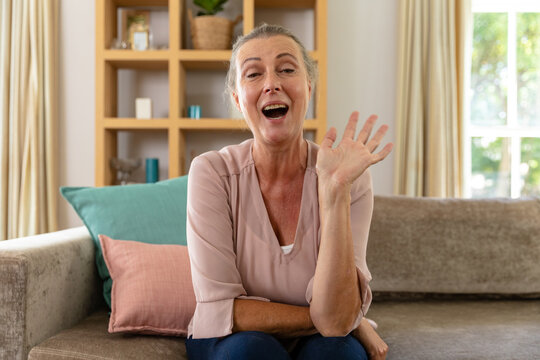 Caucasian senior woman laughing and waving hand while sitting on sofa at home - Powered by Adobe