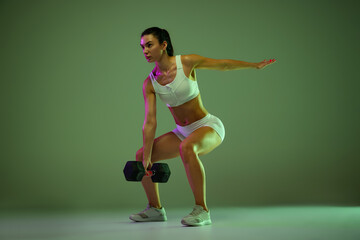 Portrait of young sportive girl doing squats, training with dumbbell isolated over green studio background in neon light