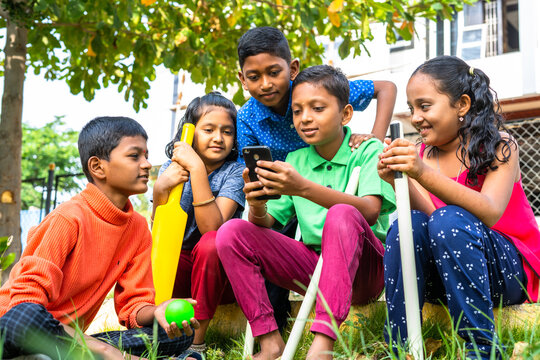 Teenage kid showing mobile phone to his friends at park after playing cricket - concept of technology, togetherness and communication