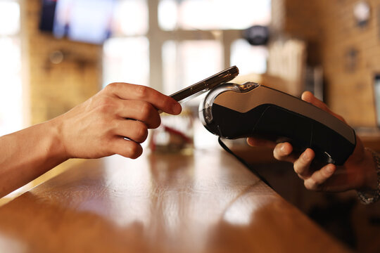Macro unknown hand giving terminal to visitor for mobile phone payment indoor. Client hand paying bill with smartphone in blurred background. Cropped shot hands making contactless transaction inside.