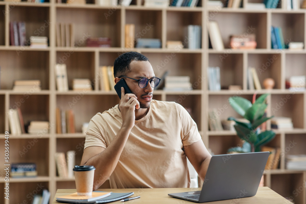 Poster remote business concept. concerned arab man talking on cellphone and using laptop while sitting at d