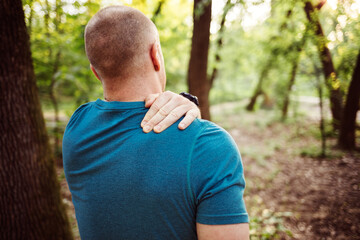 Sporty young man holding his shoulder in pain while exercising outdoors. Rear View shot of a sporty...