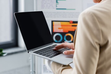 cropped view of banker using laptop with blank screen near blurred flip chart with graphs.