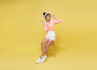 Fototapeta na wymiar Happy young Asian teen woman smiling listening music in headphones and sitting on chair isolate on bright yellow background.