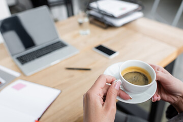 cropped view of businesswoman with coffee cup near blurred gadgets on desk.