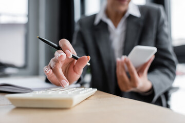 partial view of accountant working on calculator and holding smartphone on blurred background.