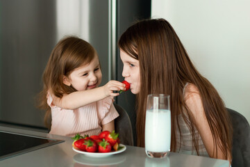 Two girls sisters sitting at kitchen, eating strawberry, drinking milk