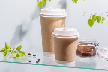 Coffee to go. Two disposable eco-friendly cardboard cups, ground coffee in a glass jar on a glass shelf on blue background with green branches and copy space. Takeaway coffee concept. Soft focus stile