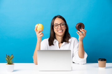 Hispanic young woman choosing between fruit and sweet chocolate donut in front of laptop in the...